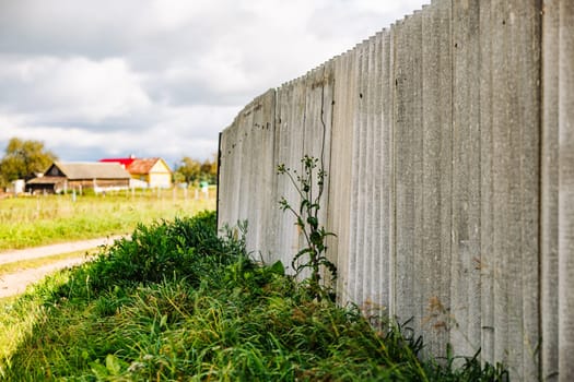 Tall grey wavy slate fence growing weeds at its base with blurred background of farm and houses
