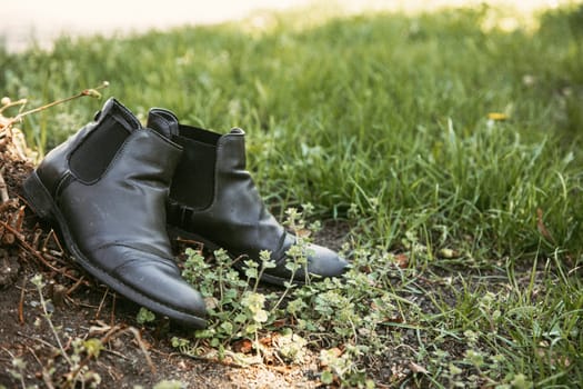 Black leather boots lying on green grass under natural sunlight. Great for projects about countryside lifestyle, walking journeys, simplicity, and forgotten objects