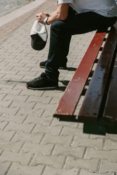 A man in jeans and sneakers sits on a bench under the sun, holding a cap with a relaxed and thoughtful posture. Perfect for lifestyle visuals, waiting scenes, or urban moments.