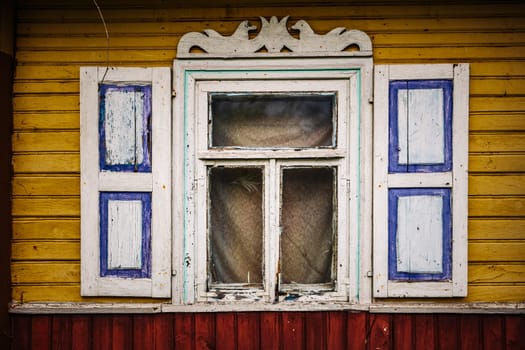Weathered window with peeling paint and decorative shutters on a yellow wooden wall, suggesting age and decay