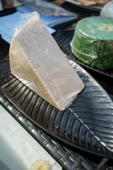 a piece of cheese in clear packaging at a farmers market. a large piece of cheese on a black oval plate.