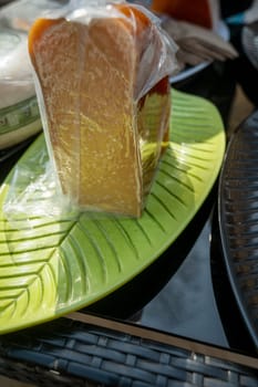 A piece of cheese, dark yellow in transparent packaging at a farmers market. Natural cheese at a city fair. A large piece of cheese on a green oval plate.