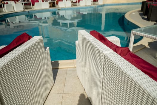 Bright blue swimming pool and dining area around it in the hotel. White wicker sofas with red pillows.