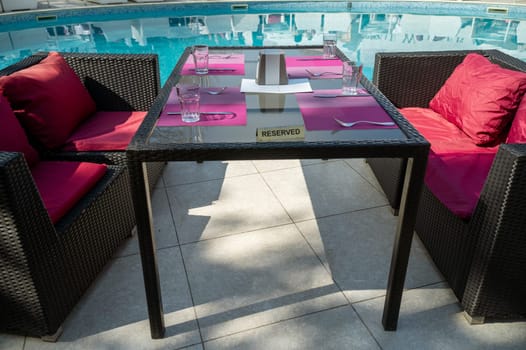 set table in hotel restaurant near swimming pool. table is set for four persons. there is a reserved sign on the table. black table with red cushions.