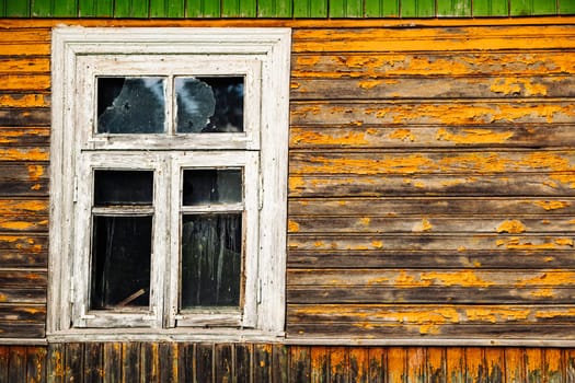 Weathered window with broken glass and peeling orange paint on a wooden facade, showing decay and neglect