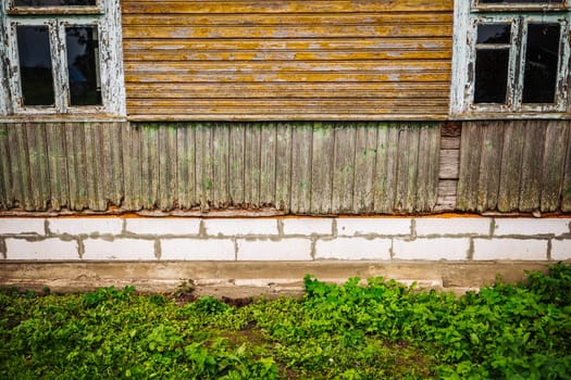 Weathered wooden wall with damaged planks and two windows showing foundation and grass