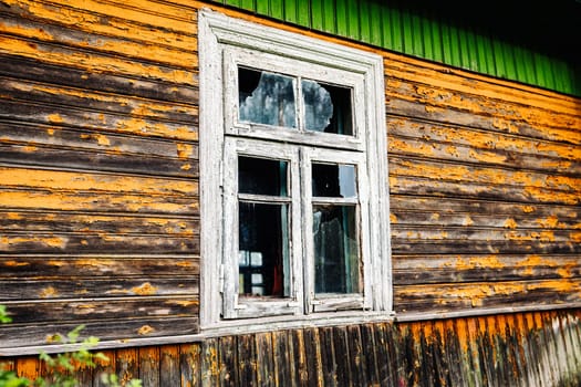 Old broken window in weathered wooden wall with peeling orange paint showing signs of decay