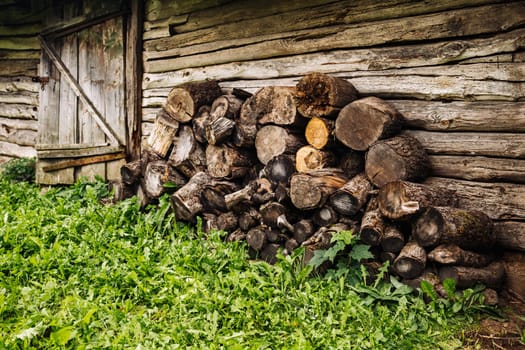 Pile of firewood logs near old wooden barn with green grass in countryside