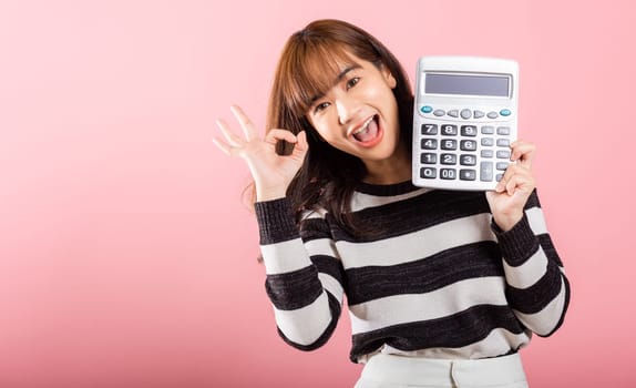 Tax day concept. Woman confident smiling holding electronic calculator and show OK gesture, excited happy Asian female studio shot isolated on pink background, Account and finance counting income