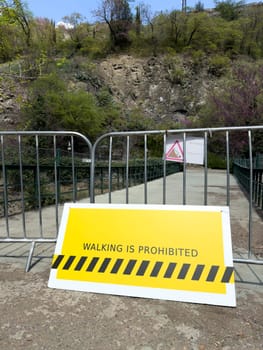 Yellow warning sign and metal barrier block access to pedestrian bridge due to danger, emphasizing safety, restricted area and walking prohibition in natural outdoor environment