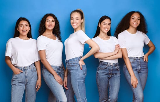 Pretty Multicultural Girls Smiling To Camera Standing On Blue Studio Background, Wearing Casual Jeans And T-Shirts. Diverse Beauty Concept.
