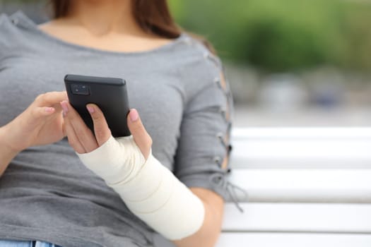 Close up portrait of a convalescent woman with bandaged arm using phone sitting on a bench in a park