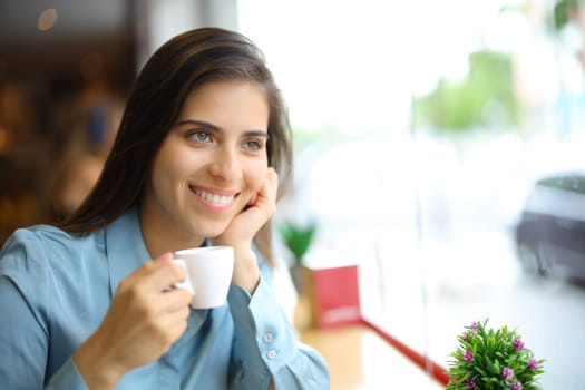 Happy woman drinking coffee in a bar looking away through a window