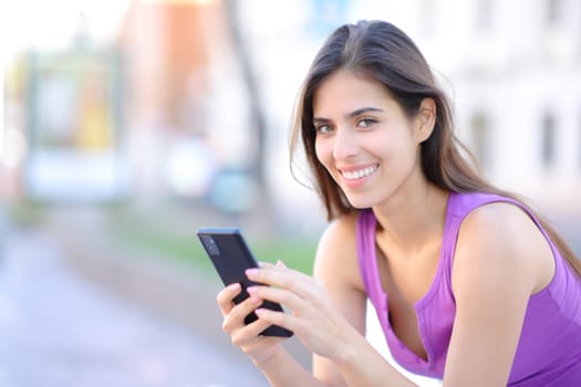 Happy phone user looks at camera sitting in the street