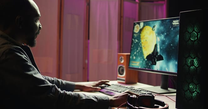 Man playing SF space videogame on PC screen in neon illuminated apartment living room. Gamer enjoying spaceship flying game on computer desktop display in RGB lit home late at night, camera B