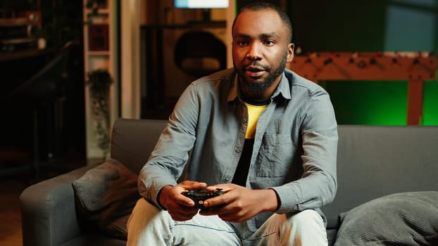 Man arriving home, gaming and enjoying leisure time. Man sitting on couch in neon illuminated living room, playing videogame on console, relaxing after hard day at work, camera A