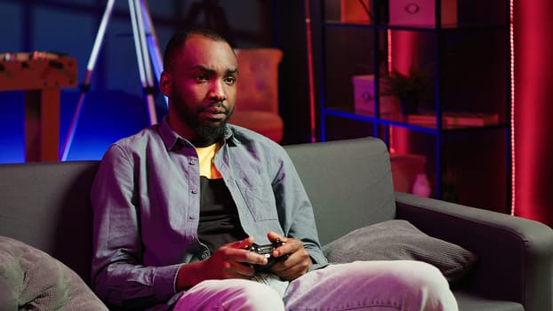Focused gamer in RGB lit home gaming with gamepad, enjoying himself. Man sitting on sofa in neon illuminated living room, playing videogames on console using controller, camera A