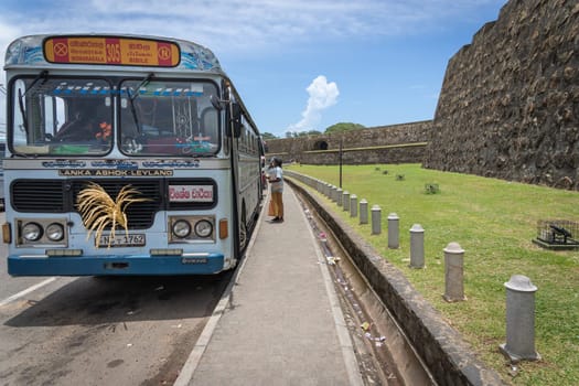 Galle Sri Lanka - September 14 2024; Decorated public transportation bus with man selling rice cakes to travelers outside the stone Galle Fort wall.