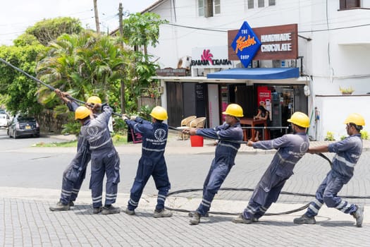 Galle Sri Lanka - September 14 2024; Workers from Ceylon Electric Power standing in row with yellow hard-hats pulling a power-line into place.