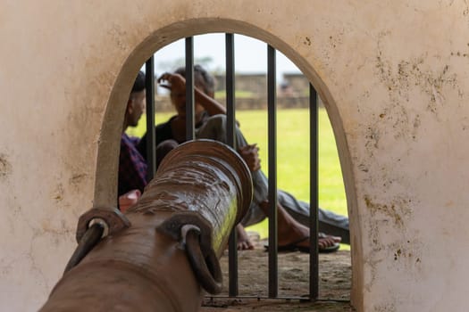 Galle Sri Lanka - September 14 2024; Ancient iron cannon pointing through grille opening in wall with blurred image of people sitting on other side.