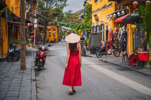 back of beautiful female tourist in traditional Vietnamese hat and red dress on street in told town in Hoi An city in Vietnam in Asia