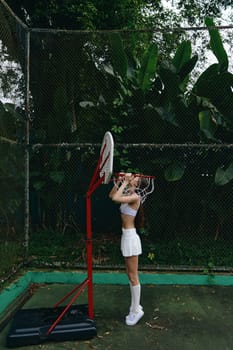 Female basketball player in a crop top and mini skirt practicing shooting on court, showcasing determination amidst tropical greenery