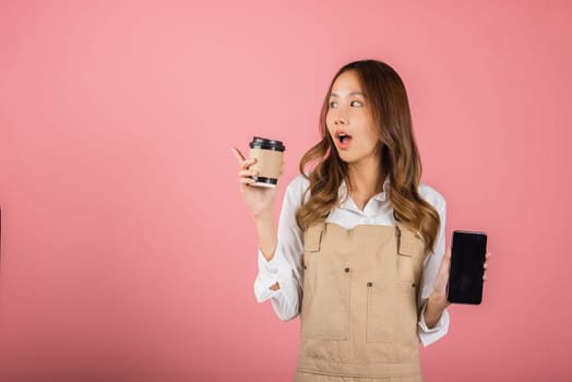 Portrait Asian happy young woman barista bar tender coffee maker holding coffee tea hot cup and mobile phone studio shot isolated on pink background, female smiling hold takeaways cup and smartphone