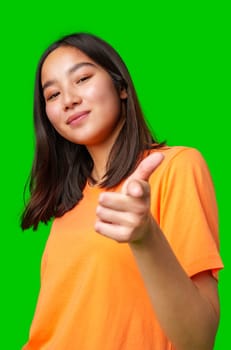 A young woman smiles confidently while pointing playfully at the camera with a bright green background. She wears an orange shirt, showcasing a lively demeanor.