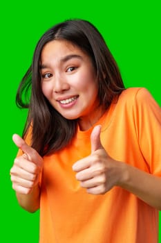 A young woman with long hair smiles broadly while giving a thumbs up with both hands. She wears an orange shirt and is set against a vivid green backdrop, portraying enthusiasm.