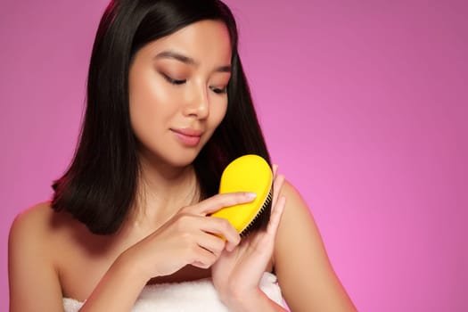 A young woman with long, black hair gently brushes her hair with a yellow brush. She appears serene and content, set against a vibrant pink backdrop that enhances her relaxed mood.