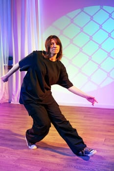 A dancer moves expressively in a black outfit, showcasing agility and skill amidst soft purple and blue lighting in a spacious studio environment.