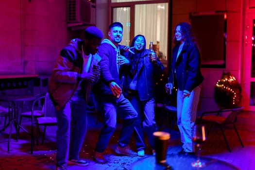 A group of friends are joyfully dancing together in a colorful outdoor space adorned with festive lights. They are celebrating and enjoying drinks in a cozy atmosphere.
