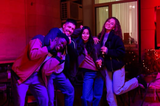 Group of four friends enjoying a lively indoor gathering at night, laughing and posing with drinks in hand under colorful lighting.