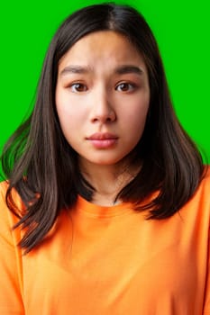 A young woman with long dark hair looks surprised while wearing an orange shirt. The bright green background creates a vibrant atmosphere, highlighting her expression.