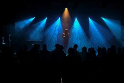 Musician stands center stage surrounded by blue lighting, creating a vibrant atmosphere. The audience watches intently, enjoying the live musical experience.