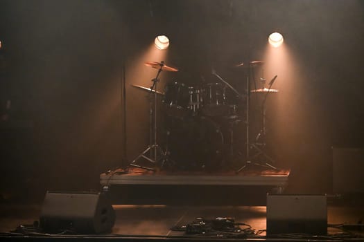 Bright stage lights illuminate the drum set as an excited crowd waits for the performance to begin, creating an atmosphere full of energy and anticipation.