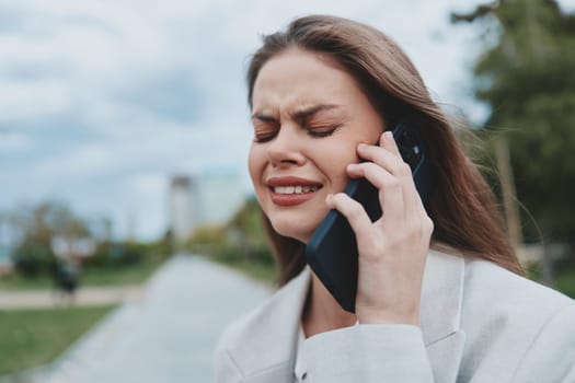 Emotional woman feeling stressed and frustrated during a phone call outdoors, wearing casual jacket with natural background, expressing anxiety and communication tension.