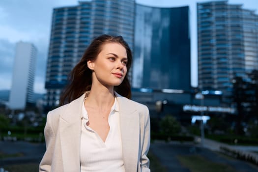 confident woman in beige blazer smiling outdoors with city buildings background, representing modern lifestyle business concept and professional fashion style