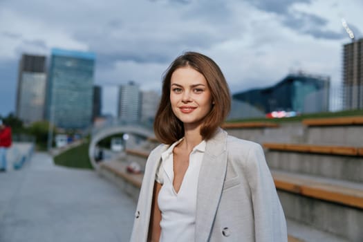 confident woman in business attire stands outdoors in urban environment with modern buildings and cloudy sky, showing casual style and professional appearance
