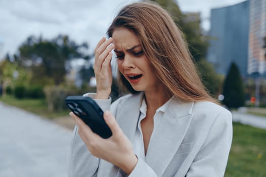 A distressed business woman in a light blazer looks at her smartphone with concern outdoors in an urban environment. Her emotion reflects the challenges of modern business.