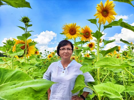 A woman stands confidently amidst tall sunflowers under a clear blue sky. The summer sun illuminates the lush green leaves, creating a joyful atmosphere in the blooming field.