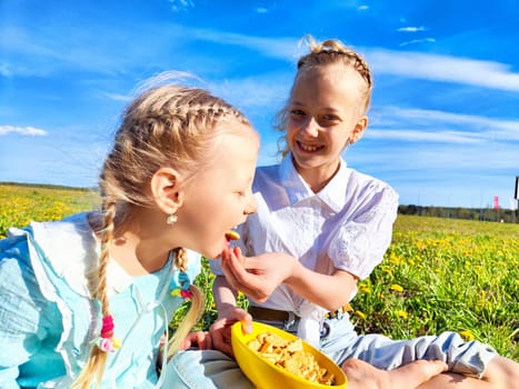Two young girls share treats in a blooming meadow under a clear blue sky on a warm spring day.
