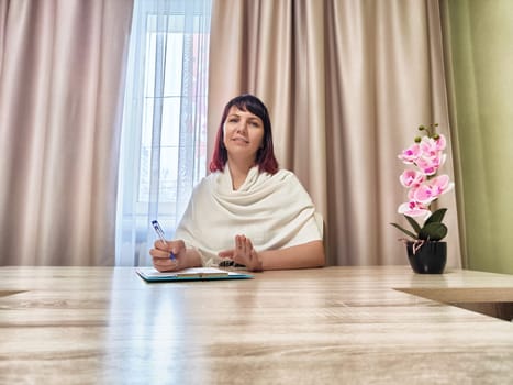 Psychologist sits at her desk, engaging with a client while taking notes, surrounded by calming colors and a plant.