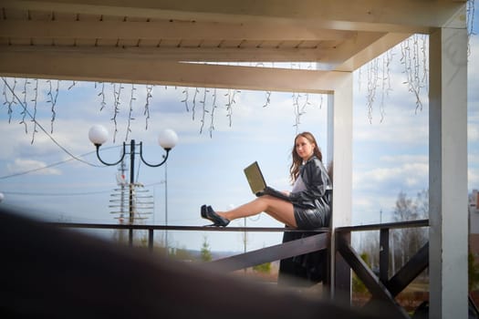 A woman focused on her laptop sits elegantly on a wooden railing in a sunny outdoor area with decorative lights.