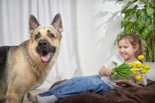 A young girl sits on a cozy surface with a bouquet of flowers while a playful German shepherd dog stands nearby, both enjoying a joyful moment.
