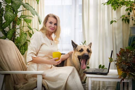 A middle-aged blonde blogger sips from a yellow mug with her German Shepherd in a cozy living space.