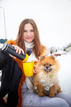 A woman serves a warm drink while her fluffy Pomeranian sits beside her in the snow, enjoying a winter outing.