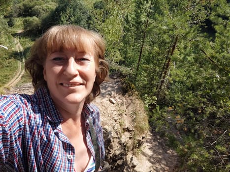 A middle-aged woman with blonde hair smiles brightly while taking a selfie on a nature trail surrounded by trees and hills, capturing her adventure.