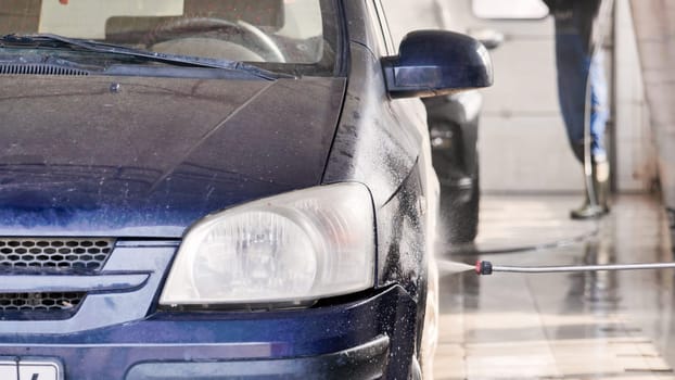 Car wash worker spraying water on dirty blue car using high pressure washer lance in service station