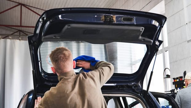 Professional car wash worker cleaning rear window of a car with open trunk, providing meticulous detailing service